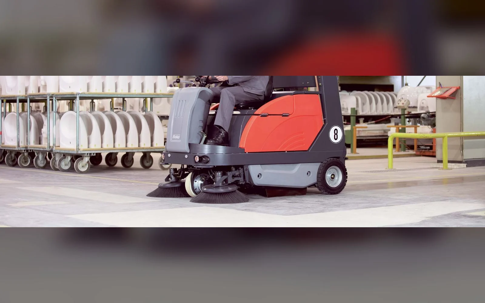 Operator driving a Hako Sweepmaster ride-on floor sweeper in a production hall